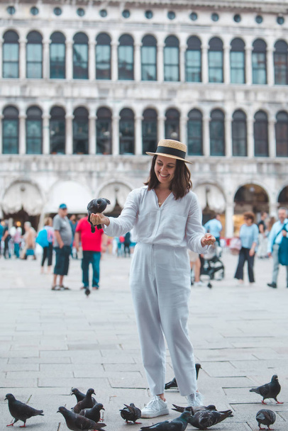 kobieta w białych ubraniach ze słomkowym kapeluszem zabawia się z gołębiami na placu miejskim w Wenecji - Zdjęcie, obraz