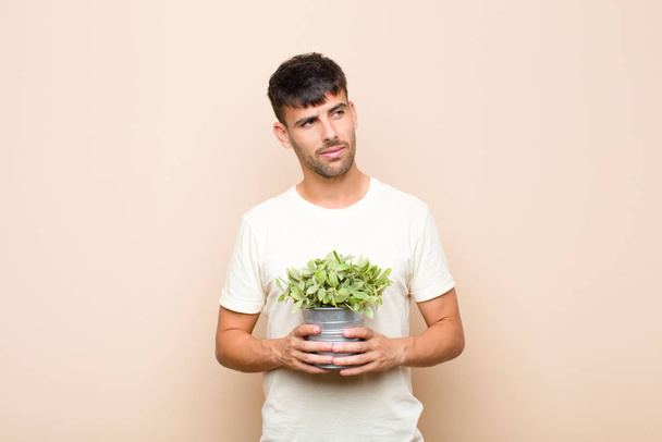 young handsome man feeling sad, upset or angry and looking to the side with a negative attitude, frowning in disagreement holding a plant - Valokuva, kuva