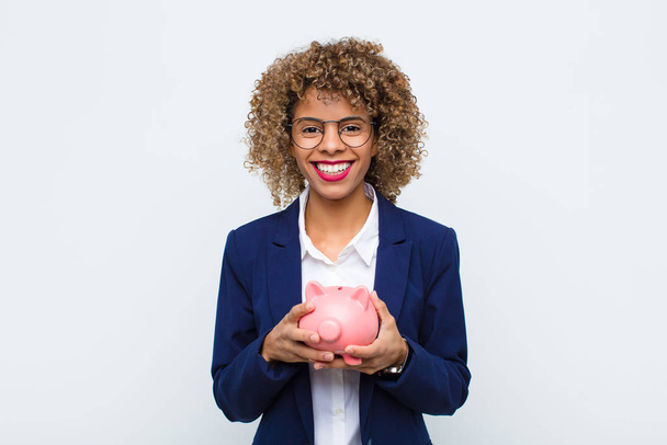 young african american woman smiling happily with a hand on hip and confident, positive, proud and friendly attitude with a piggy bank - Photo, image