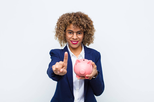young african american woman smiling proudly and confidently making number one pose triumphantly, feeling like a leader with a piggy bank - Fotó, kép