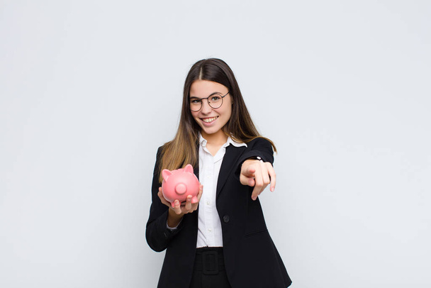 young pretty woman pointing at camera with a satisfied, confident, friendly smile, choosing you with a piggy bank - Photo, image