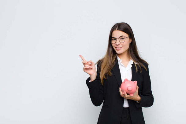 young pretty woman smiling cheerfully, feeling happy and pointing to the side and upwards, showing object in copy space with a piggy bank - Foto, Imagen