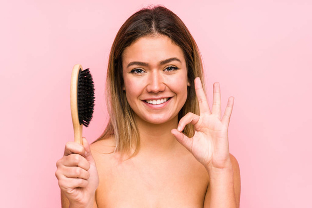 Jovem caucasiana segurando uma escova de cabelo isolado alegre e confiante mostrando ok gesto
. - Foto, Imagem