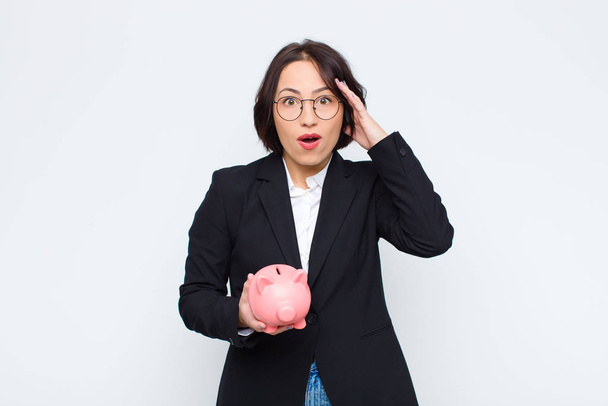 young pretty woman looking happy, astonished and surprised, smiling and realizing amazing and incredible good news with a piggy bank - Photo, image