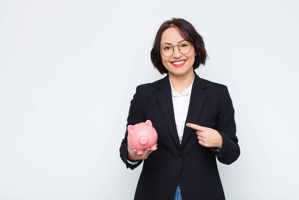 young pretty woman smiling cheerfully, feeling happy and pointing to the side and upwards, showing object in copy space with a piggy bank - Photo, Image