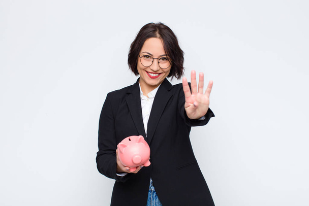 young pretty woman smiling and looking friendly, showing number four or fourth with hand forward, counting down with a piggy bank - Photo, image
