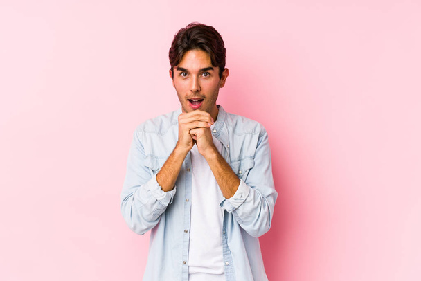 Jeune homme caucasien posant dans un fond rose isolé priant pour la chance, étonné et ouvrant la bouche regardant vers l'avant. - Photo, image
