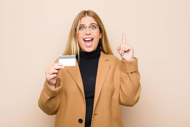 young blonde woman feeling like a happy and excited genius after realizing an idea, cheerfully raising finger, eureka! with a credit card - Foto, imagen
