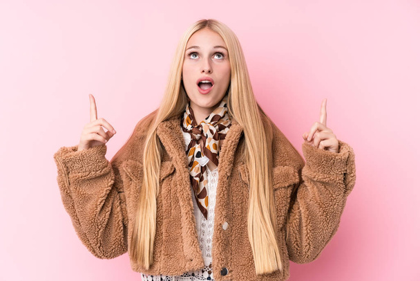 Nuori blondi nainen yllään takki vastaan vaaleanpunainen tausta osoittaa ylösalaisin avattu suu
. - Valokuva, kuva