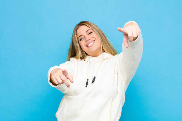 young blonde woman feeling happy and confident, pointing to camera with both hands and laughing, choosing you against flat wall - Zdjęcie, obraz