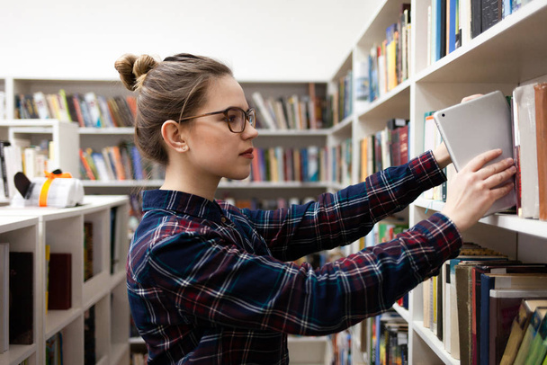 Ein junger Student stellt ein Tablet in ein Regal mit Büchern in der Bibliothek. Konzept moderner Lesemethoden - Foto, Bild