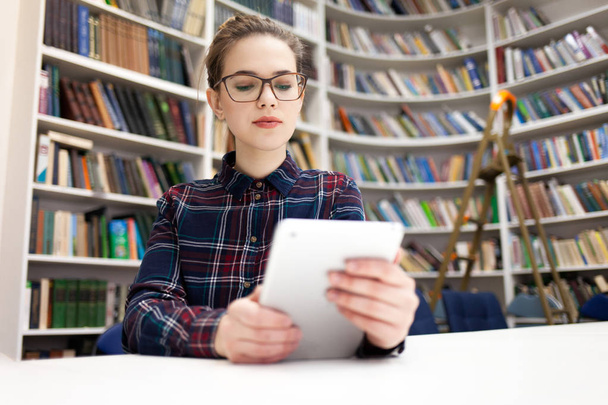 Egy lány, aki tabletekkel dolgozik a könyvtárban. A diák jobban szeret táblagéppel dolgozni, mint könyvekkel. - Fotó, kép