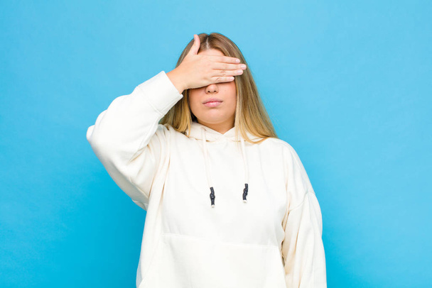 young blonde woman covering eyes with one hand feeling scared or anxious, wondering or blindly waiting for a surprise against flat wall - Foto, imagen