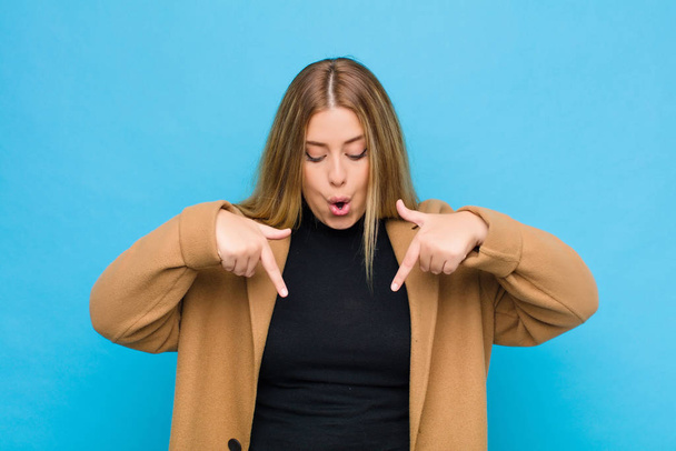 young blonde woman feeling shocked, open-mouthed and amazed, looking and pointing downwards in disbelief and surprise against flat wall - Foto, Bild