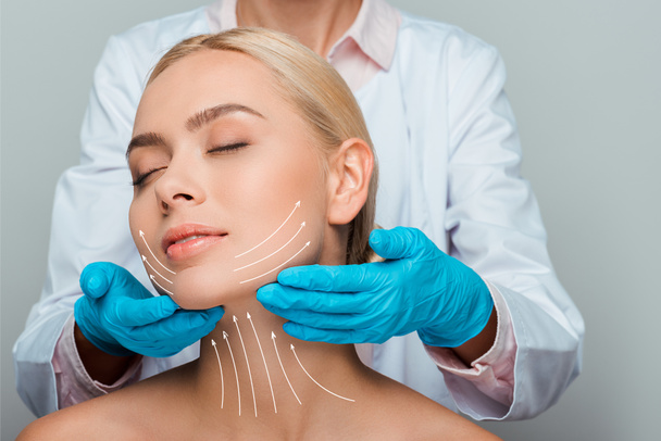 cropped view of beautician in blue latex gloves touching neck of naked woman with closed eyes and facelift marks on face isolated on grey  - Photo, Image