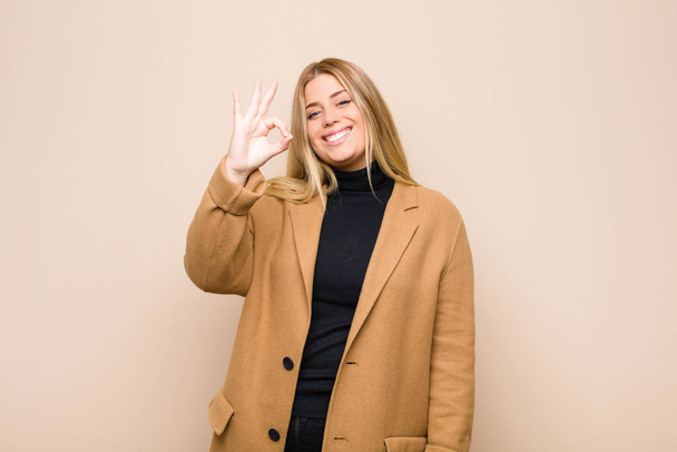 young blonde woman feeling happy, relaxed and satisfied, showing approval with okay gesture, smiling against flat wall - Fotó, kép