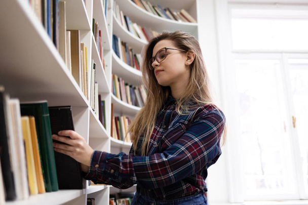 Cute student in glasses takes a book from the library bookshelf. Young lady is looking for the right literature among many books - Фото, зображення