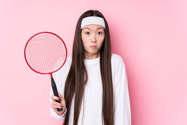 Young chinese woman playing badminton isolated shrugs shoulders and open eyes confused. - Photo, Image