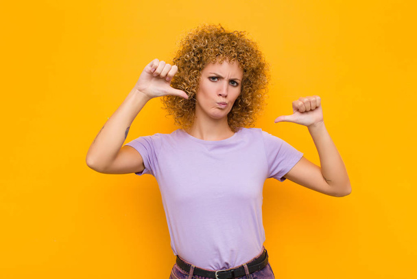 young afro woman looking sad, disappointed or angry, showing thumbs down in disagreement, feeling frustrated against orange wall - Foto, Imagem