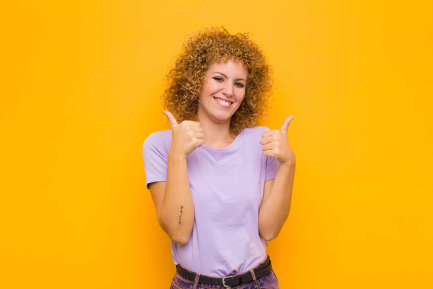 young afro woman smiling joyfully and looking happy, feeling carefree and positive with both thumbs up against orange wall - Photo, Image