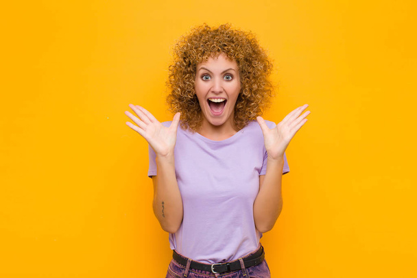 young afro woman feeling happy, excited, surprised or shocked, smiling and astonished at something unbelievable against orange wall - Photo, image