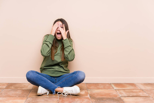 Молода біла жінка сидить на підлозі ізольовано блимає крізь пальці переляканими і нервовими
. - Фото, зображення