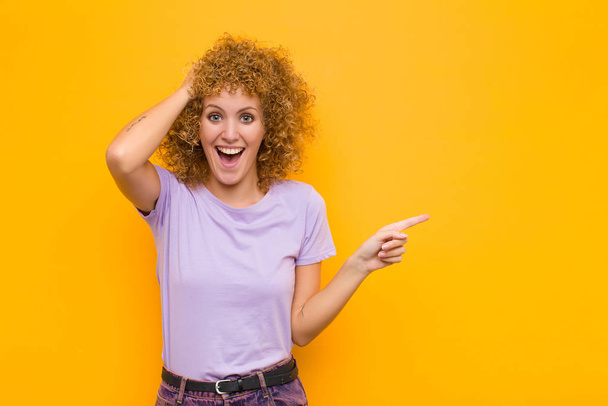 young afro woman laughing, looking happy, positive and surprised, realizing a great idea pointing to lateral copy space against orange wall - Photo, image