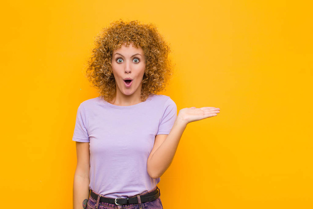 young afro woman looking surprised and shocked, with jaw dropped holding an object with an open hand on the side against orange wall - Foto, Bild