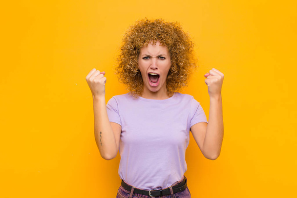 young afro woman shouting aggressively with an angry expression or with fists clenched celebrating success against orange wall - Photo, image