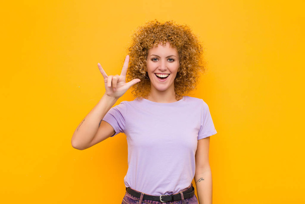 young afro woman feeling happy, fun, confident, positive and rebellious, making rock or heavy metal sign with hand against orange wall - Fotó, kép