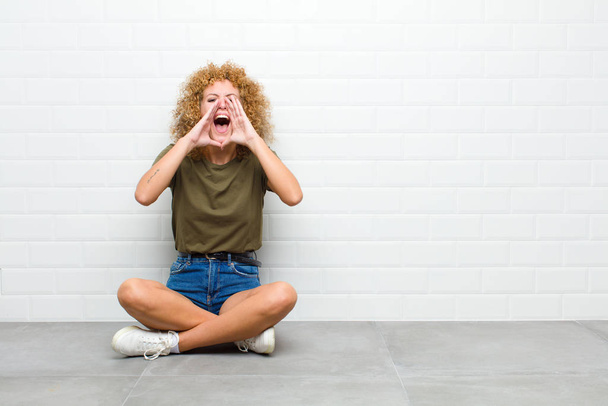 young afro woman feeling happy, excited and positive, giving a big shout out with hands next to mouth, calling out sitting on a floor - Foto, Imagen