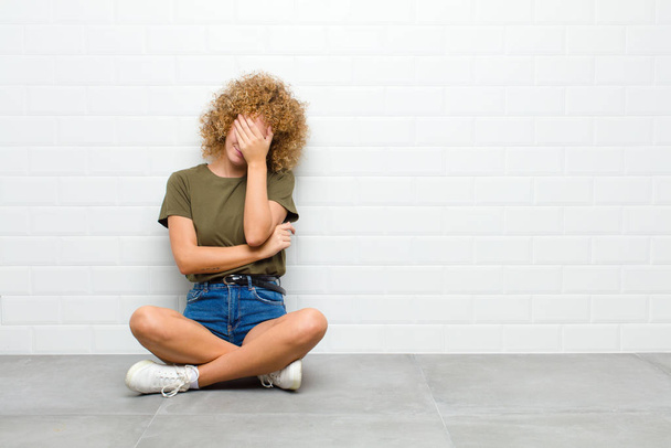 young afro woman looking stressed, ashamed or upset, with a headache, covering face with hand sitting on a floor - Foto, imagen