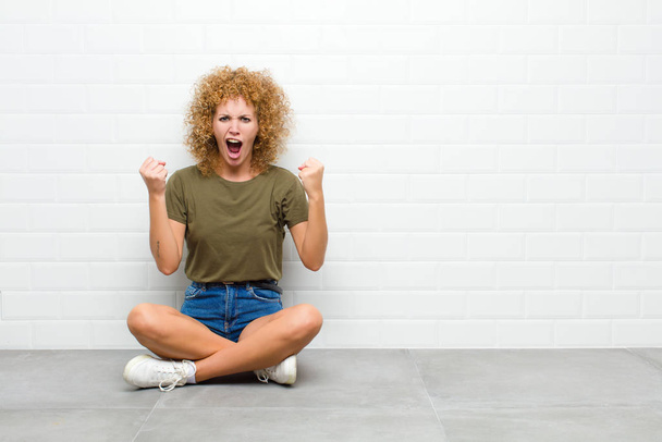young afro woman shouting aggressively with an angry expression or with fists clenched celebrating success sitting on a floor - Фото, изображение