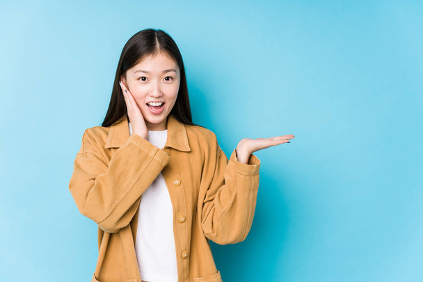 Young chinese woman posing in a blue background isolated holds copy space on a palm, keep hand over cheek. Amazed and delighted. - Photo, Image