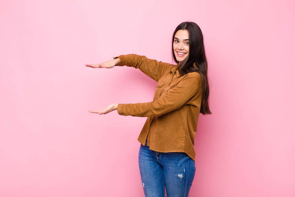 young pretty woman smiling, feeling happy, positive and satisfied, holding or showing object or concept on copy space against pink wall - Фото, зображення