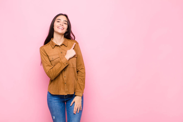 young pretty woman smiling cheerfully, feeling happy and pointing to the side and upwards, showing object in copy space against pink wall - Fotoğraf, Görsel
