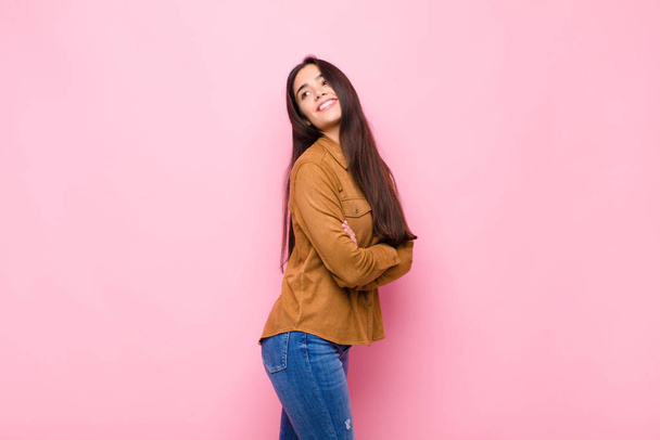 young pretty woman smiling gleefully, feeling happy, satisfied and relaxed, with crossed arms and looking to the side against pink wall - Zdjęcie, obraz
