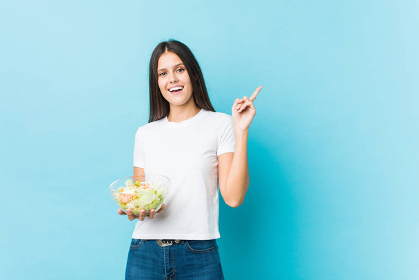 Joven mujer caucásica sosteniendo una ensalada sonriendo alegremente señalando con el dedo índice de distancia
. - Foto, Imagen