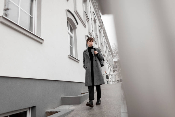 シックなグレーのコートを着たヨーロッパの若い男性は、革のファッショナブルなバックパックを備えた黒いスタイリッシュなブーツで、壁の近くに屋外で散歩しています。上品な服を着たトレンディ魅力的な現代人。ユース・スタイル. - 写真・画像