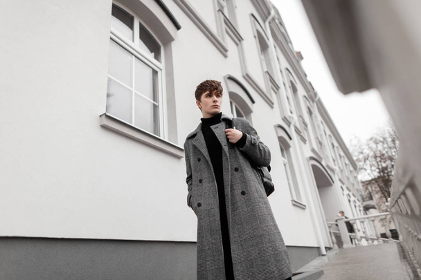 Νεαρό δροσερό άνθρωπος μοντέλο σε ένα χειμωνιάτικο καρό παλτό με ένα κομψό μαύρο δερμάτινο σακίδιο βόλτες στο δρόμο κοντά σε ένα κτίριο. Αστικός όμορφος άντρας με κομψά ρούχα έξω. Νέα ανδρικά ενδύματα συλλογής μόδας. - Φωτογραφία, εικόνα