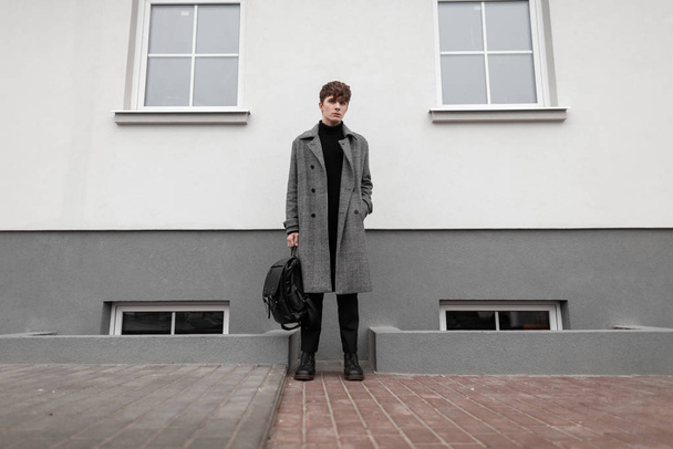 Fashionable young man in a gray elegant plaid coat with a leather stylish black backpack in autumn trendy boots stands near a vintage wall. Handsome modern guy in the city. Youth seasonal menswear.  - Foto, Bild