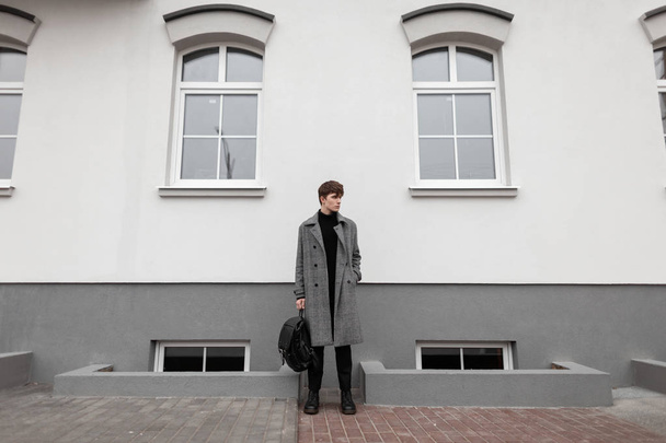 Trendy jongeman in een vintage geruite grijze jas in zwarte stijlvolle laarzen met een leren modieuze rugzak staat buiten bij de muur. Aantrekkelijke moderne man in elegante kleren. Jongerenstijl. - Foto, afbeelding