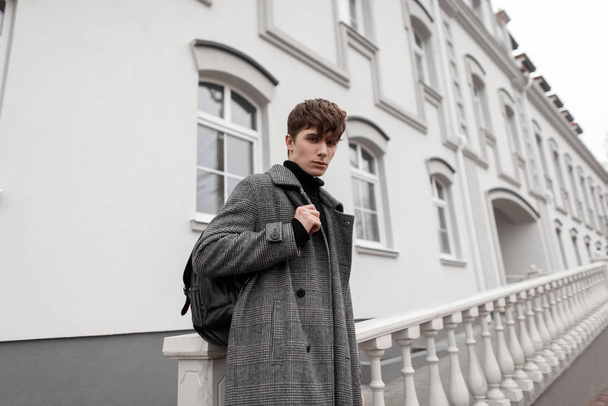 Κομψό όμορφος νεαρός άνδρας σε ένα μοντέρνο καρό παλτό με ένα κομψό δερμάτινο σακίδιο με ένα μοντέρνο χτένισμα στέκεται στο δρόμο κοντά σε ένα κτίριο σε μια ημέρα του φθινοπώρου. Εποχιακά εξωτερικά ενδύματα σύγχρονων ανδρών. - Φωτογραφία, εικόνα