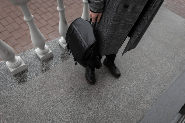 Een close-up. Modieuze seizoensgebonden buitenkleding voor mannen. De Europese jonge urban man staat in een stijlvolle geruite grijze jas in klassieke zwarte broek in vintage laarzen met een stoere leren rugzak. Mode. - Foto, afbeelding