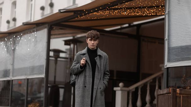 Attractive stylish young man in fashionable winter clothes with a trendy backpack stands in the city near a cafe decorated with New Year vintage lights. Modern urban guy on the street. Youth style. - 写真・画像