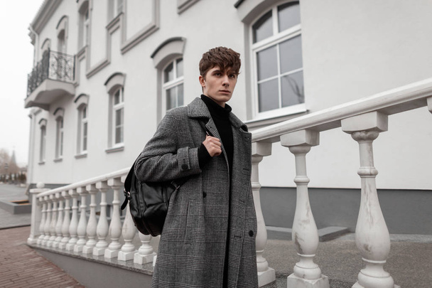 Европейский молодой человек в модном сером клетчатом пальто со стильной прической с кожаным черным рюкзаком стоит в городе рядом со старинным зданием. Attractive guy is a fashion model.Fashionable men swear
 - Фото, изображение