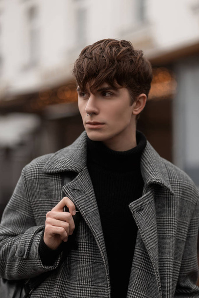 Fashion model of a young attractive man in a warm black shirt in a fashionable plaid coat with a leather backpack with a trendy hairstyle. Portrait of a handsome guy in elegant autumn outerwear. - Valokuva, kuva