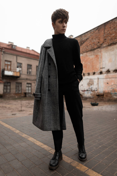 Νεαρό ευρωπαϊκό ανδρικό μοντέλο με μοντέρνο χτένισμα σε ένα κομψό γκρι καρό παλτό σε τζιν με μαύρο vintage πουλόβερ στέκεται στο δρόμο ανάμεσα στα κτίρια. Όμορφος μοντέρνος Ευρωπαίος.. - Φωτογραφία, εικόνα