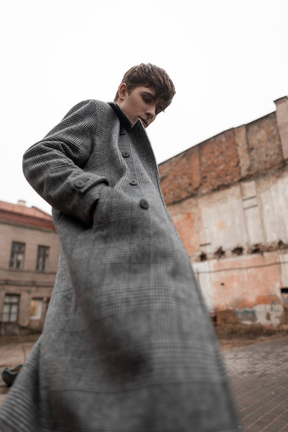 In a fashionable gray plaid long coat a handsome stylish young man with a trendy hairstyle is standing on the street. Attractive guy model in seasonal clothes poses outdoors. New collection menswear. - Foto, Bild