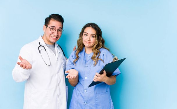 Young doctor couple posing in a blue background isolated doubting and shrugging shoulders in questioning gesture. - Fotoğraf, Görsel